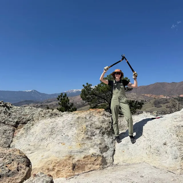Volunteer holding loppers overhead on a sandstone outcrop with mountains behind